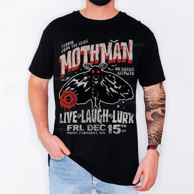 May include: A black t-shirt featuring a graphic of a moth with red eyes. The text reads: TERROR FROM THE SKIES, MOTHMAN, NO SISSIES ALLOWED, LIVE LAUGH LURK, FRI. DEC 15TH 67, POINT PLEASANT, WV. A red circular stamp says GUARANTEED TO SCARE.