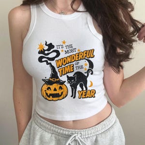 May include: White tank top with a Halloween-themed graphic. The design includes a jack-o'-lantern wearing a witch hat, a black cat, and the text "It's the Most Wonderful Time of the Year."