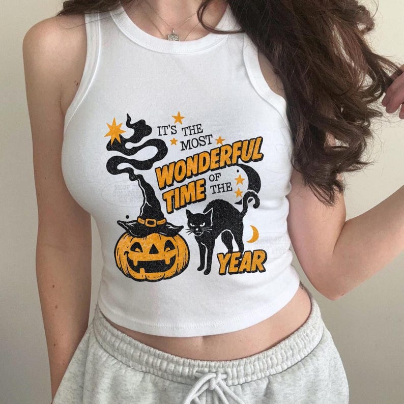 May include: White tank top with a Halloween-themed graphic. The design includes a jack-o'-lantern wearing a witch hat, a black cat, and the text "It's the Most Wonderful Time of the Year."
