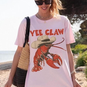 Op de afbeelding: Lichtroze T-shirt met een rode kreeft met een cowboyhoed en de tekst "YEE CLAW" in het rood. De persoon draagt een strooien tas en bevindt zich op het strand.