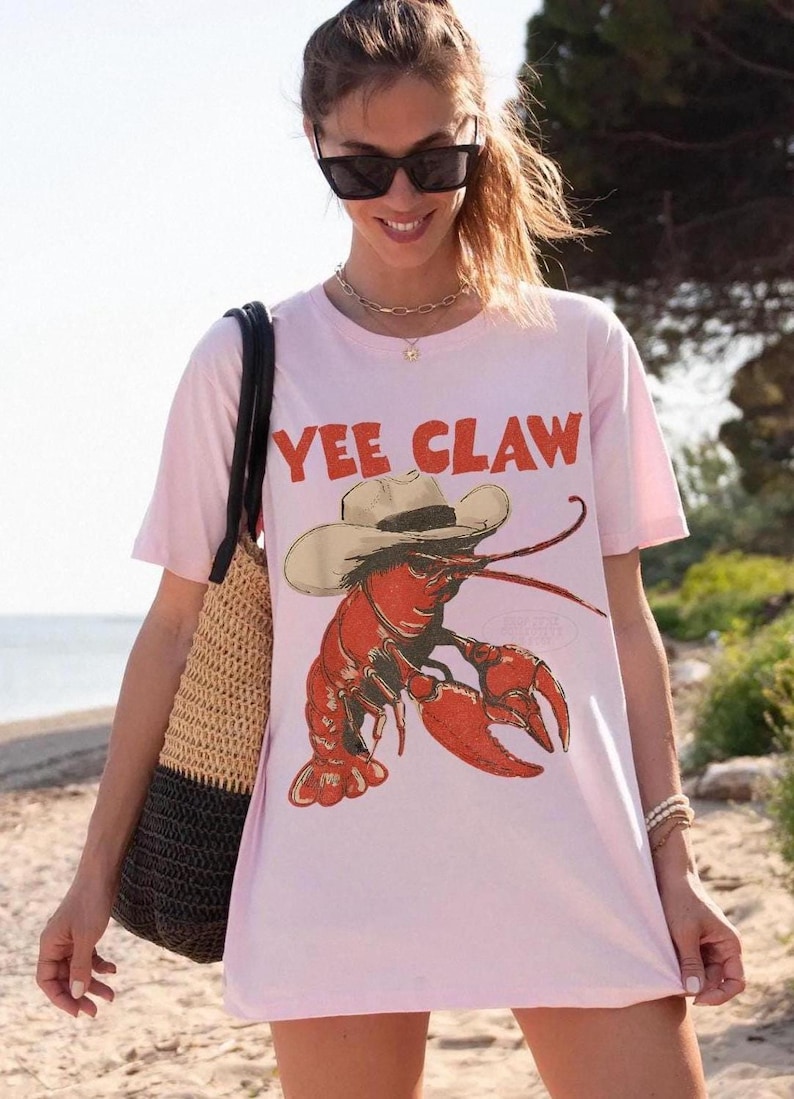 Op de afbeelding: Lichtroze T-shirt met een rode kreeft met een cowboyhoed en de tekst "YEE CLAW" in het rood. De persoon draagt een strooien tas en bevindt zich op het strand.