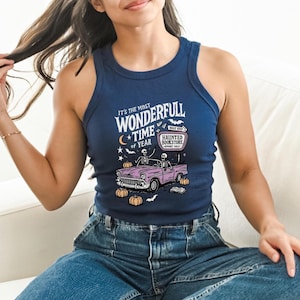 May include: Navy blue tank top with a Halloween-themed graphic. The graphic features a pink car with skeletons, pumpkins, and the text "It's the most wonderfull time of year" and "Haunted Bookstore".
