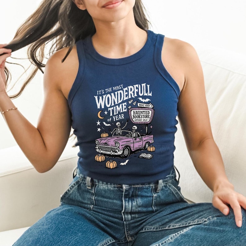May include: Navy blue tank top with a Halloween-themed graphic. The graphic features a pink car with skeletons, pumpkins, and the text "It's the most wonderfull time of year" and "Haunted Bookstore".