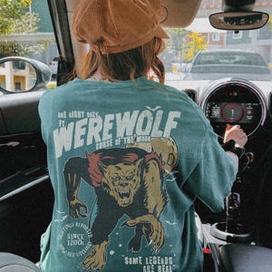 May include: A teal t-shirt featuring a graphic of a snarling werewolf and a full moon. The text reads: One Night Only ft. Werewolf Curse of the Moon, Sightings Reported Since 1200s Now See Upclose, and Some Legends Are Real. The shirt is worn with a brown hat.