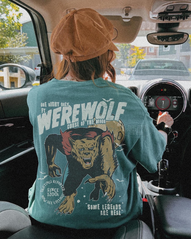 May include: A teal t-shirt featuring a graphic of a snarling werewolf and a full moon. The text reads: One Night Only ft. Werewolf Curse of the Moon, Sightings Reported Since 1200s Now See Upclose, and Some Legends Are Real. The shirt is worn with a brown hat.