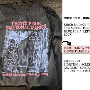 Peut inclure: Un t-shirt gris anthracite avec une impression graphique en rose saumon et bleu crayeux. Le motif repr&eacute;sente une chauve-souris, un sasquatch, des fant&ocirc;mes et un cerf, avec le texte "PROTECT OUR NATIONAL PARKS" et "WHERE LEGENDS STILL ROAM FREE".
