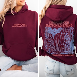 May include: A maroon hooded sweatshirt featuring the text "PROTECT OUR NATIONAL PARKS" on the front. The back displays a blue graphic illustration of a forest with a bat, a sasquatch, ghosts, and a deer, with the text "PROTECT OUR NATIONAL PARKS" and "LEGENDS STILL ROAM FREE."