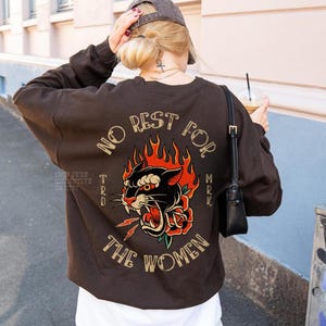May include: A brown sweatshirt with a graphic on the back featuring a black panther, red rose, and flames. The text reads "NO REST FOR THE WOMEN". The sweatshirt is worn with a white shirt and a gray cap.