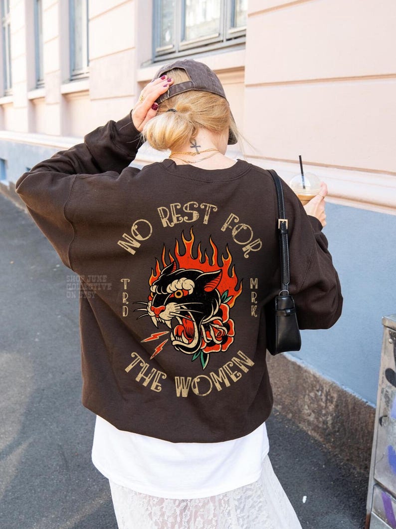 May include: A brown sweatshirt with a graphic on the back featuring a black panther, red rose, and flames. The text reads "NO REST FOR THE WOMEN". The sweatshirt is worn with a white shirt and a gray cap.