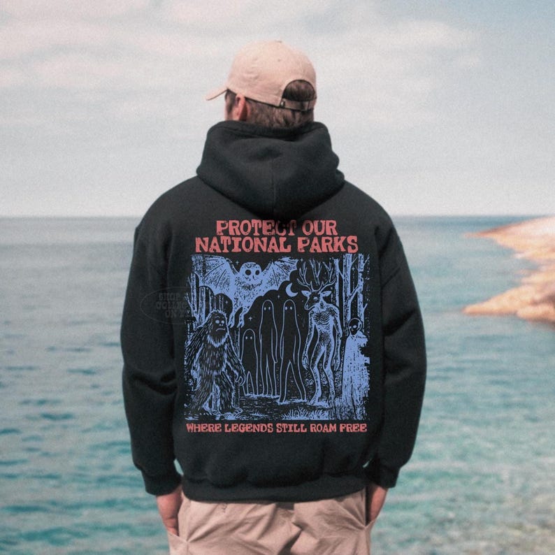 May include: A black hooded sweatshirt featuring a graphic print on the back with the text PROTECT OUR NATIONAL PARKS and WHERE LEGENDS STILL ROAM FREE. The illustration depicts a forest scene with a sasquatch, an owl, and other figures.