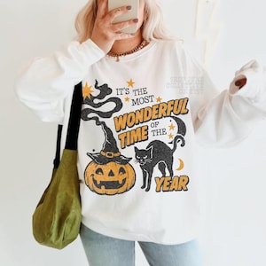 Peut inclure: Un sweat-shirt blanc &agrave; manches longues avec une impression graphique portant le texte IT'S THE MOST WONDERFUL TIME OF THE YEAR. Le motif comprend un chapeau de sorci&egrave;re noir sur une citrouille orange, un chat noir, des &eacute;toiles et une lune.