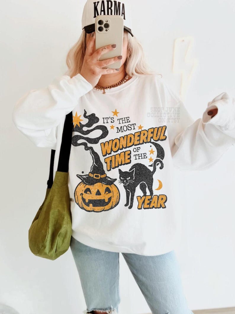 Peut inclure: Un sweat-shirt blanc &agrave; manches longues avec une impression graphique portant le texte IT'S THE MOST WONDERFUL TIME OF THE YEAR. Le motif comprend un chapeau de sorci&egrave;re noir sur une citrouille orange, un chat noir, des &eacute;toiles et une lune.