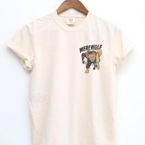 May include: A cream-colored short-sleeve t-shirt featuring a graphic on the left chest. The graphic depicts a snarling werewolf with brown fur and a red collar, positioned below the black text that reads WEREWOLF in a bold, stylized font.