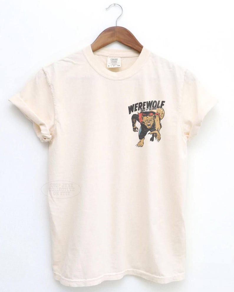 May include: A cream-colored short-sleeve t-shirt featuring a graphic on the left chest. The graphic depicts a snarling werewolf with brown fur and a red collar, positioned below the black text that reads WEREWOLF in a bold, stylized font.