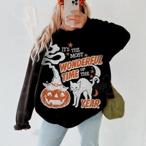 Peut inclure: Un sweat-shirt noir &agrave; manches longues avec une impression graphique portant le texte "IT'S THE MOST WONDERFUL TIME OF THE YEAR" en orange et blanc. Le motif comprend un chat blanc, une citrouille orange avec un chapeau de sorci&egrave;re et des &eacute;toiles.