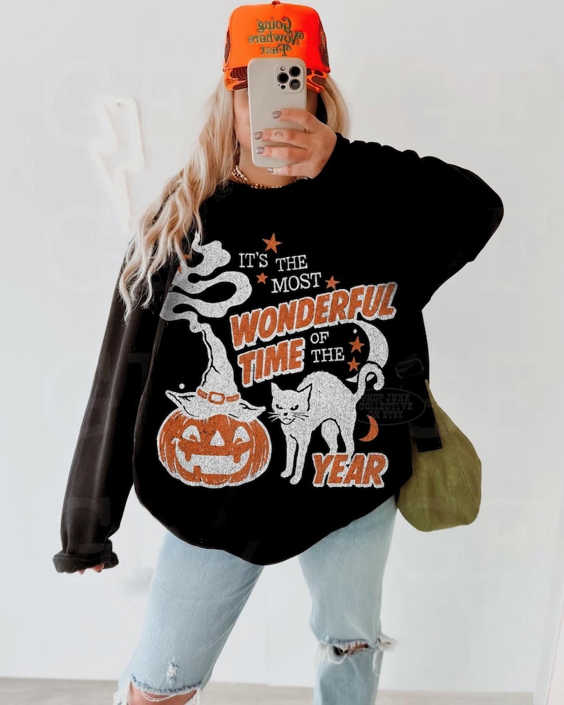 Peut inclure: Un sweat-shirt noir &agrave; manches longues avec une impression graphique portant le texte "IT'S THE MOST WONDERFUL TIME OF THE YEAR" en orange et blanc. Le motif comprend un chat blanc, une citrouille orange avec un chapeau de sorci&egrave;re et des &eacute;toiles.