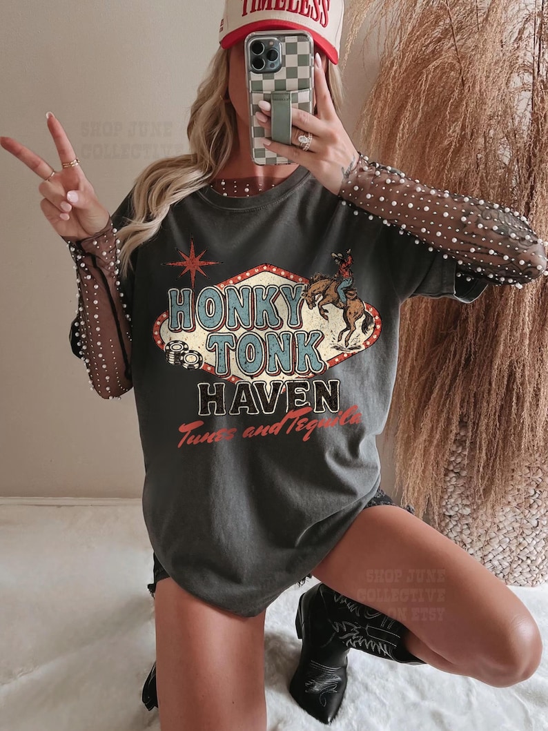 Pode incluir: T-shirt cinza escuro com o texto "Honky Tonk Haven Tunes and Tequila" em um design retr&ocirc;. A camisa apresenta um gr&aacute;fico de um cowboy montando um cavalo e uma estrela vermelha. As mangas s&atilde;o transparentes com bolinhas brancas.