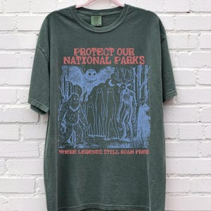 Peut inclure: Un t-shirt &agrave; manches courtes vert fonc&eacute; avec une impression graphique en rouge et bleu clair. Le texte indique PROTECT OUR NATIONAL PARKS et WHERE LEGENDS STILL ROAM FREE. L'illustration repr&eacute;sente un hibou, un sasquatch, des fant&ocirc;mes et un cerf.