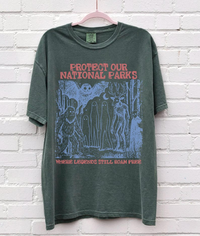 Peut inclure: Un t-shirt &agrave; manches courtes vert fonc&eacute; avec une impression graphique en rouge et bleu clair. Le texte indique PROTECT OUR NATIONAL PARKS et WHERE LEGENDS STILL ROAM FREE. L'illustration repr&eacute;sente un hibou, un sasquatch, des fant&ocirc;mes et un cerf.