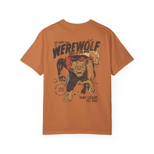 May include: A burnt orange short-sleeve t-shirt featuring a graphic on the back of a snarling werewolf. The text reads "ONE NIGHT ONLY", "WEREWOLF", "CURSE OF THE MOON", "SIGHTINGS REPORTED SINCE 1200", "NOW SEE LIKENESS", and "SOME LEGENDS ARE REAL".