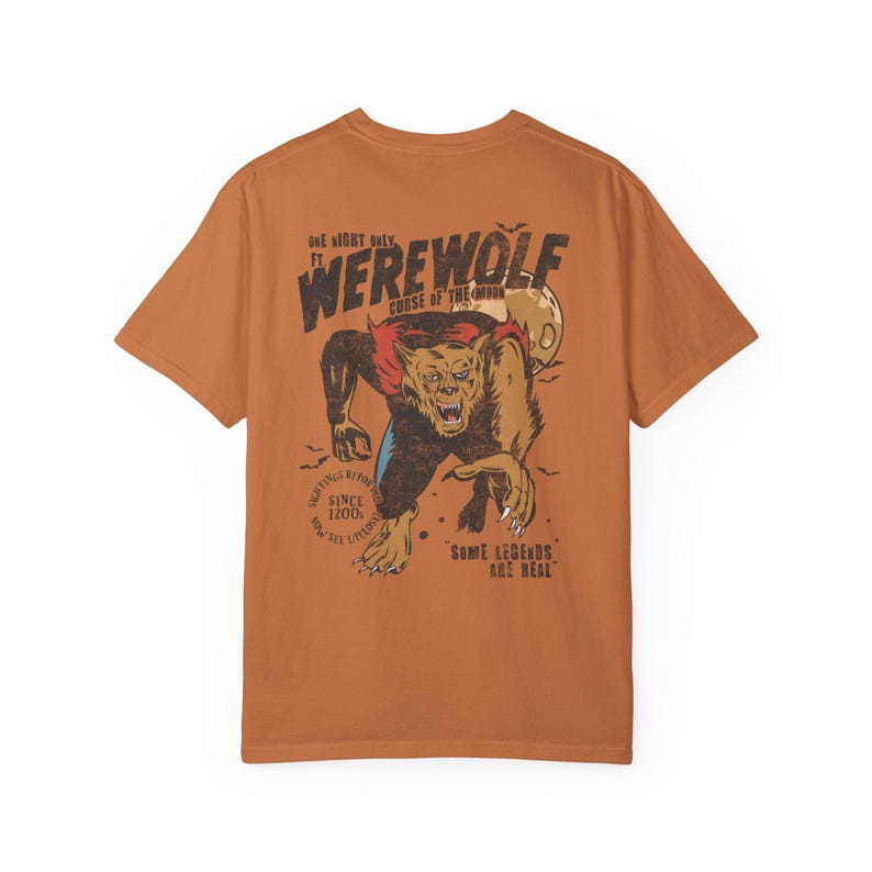 May include: A burnt orange short-sleeve t-shirt featuring a graphic on the back of a snarling werewolf. The text reads "ONE NIGHT ONLY", "WEREWOLF", "CURSE OF THE MOON", "SIGHTINGS REPORTED SINCE 1200", "NOW SEE LIKENESS", and "SOME LEGENDS ARE REAL".