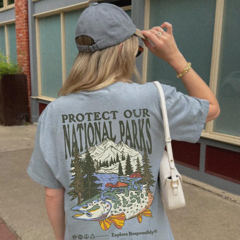 May include: A light blue t-shirt with a graphic print of a trout, mountains, and trees. The text "Protect Our National Parks" is printed on the shirt. The text "Explore Responsibly" is printed at the bottom of the shirt.