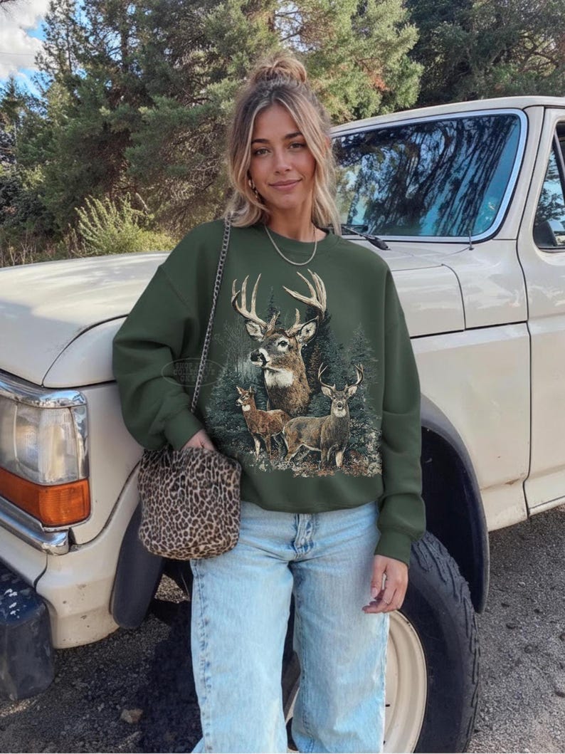 May include: An olive green sweatshirt featuring a detailed graphic of a deer family in a forest setting. The sweatshirt is paired with light blue jeans and a leopard print purse. The background includes a vintage white truck and trees.