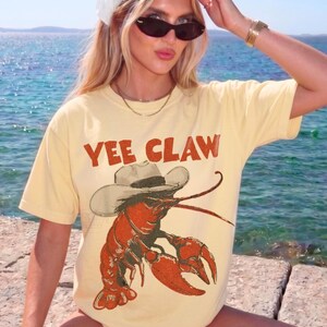 Op de afbeelding: Lichtgeel T-shirt met de tekst "YEE CLAW" in rode letters. Het shirt heeft een cartoon illustratie van een rode kreeft met een cowboyhoed. Het shirt is een casual stijl.