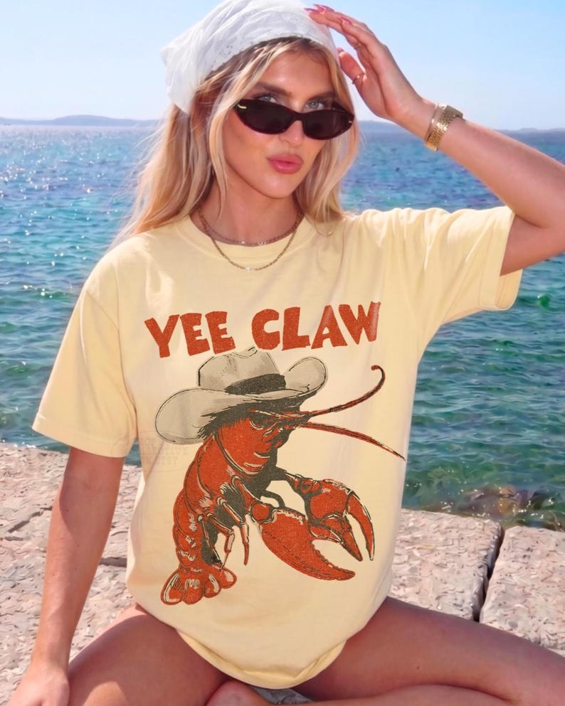 Op de afbeelding: Lichtgeel T-shirt met de tekst "YEE CLAW" in rode letters. Het shirt heeft een cartoon illustratie van een rode kreeft met een cowboyhoed. Het shirt is een casual stijl.