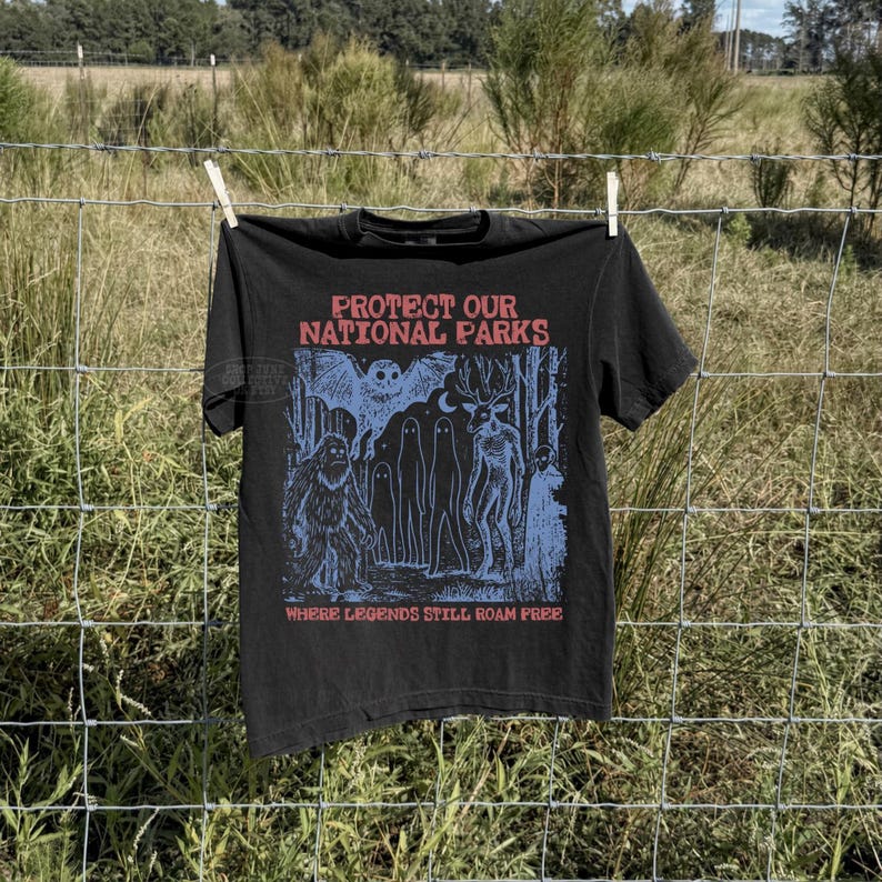 May include: A black t-shirt featuring a blue graphic print of a forest scene with a bat, a sasquatch, ghosts, and a deer-like creature. The text reads "PROTECT OUR NATIONAL PARKS" at the top and "WHERE LEGENDS STILL ROAM FREE" at the bottom in red.