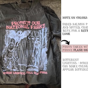 May include: A charcoal gray t-shirt featuring a graphic print in salmon pink and chalky blue. The design includes a bat, a sasquatch, ghosts, and a deer, with the text "PROTECT OUR NATIONAL PARKS" and "WHERE LEGENDS STILL ROAM FREE".
