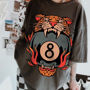 May include: Dark gray t-shirt featuring a graphic of a snarling leopard head with orange and black spots, biting a gray eight-ball with the number 8. Flames in orange and red surround the eight-ball. The shirt is a casual, oversized fit.