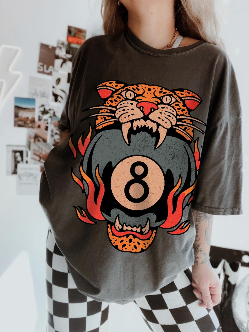 May include: Dark gray t-shirt featuring a graphic of a snarling leopard head with orange and black spots, biting a gray eight-ball with the number 8. Flames in orange and red surround the eight-ball. The shirt is a casual, oversized fit.