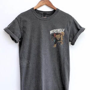 May include: A charcoal gray short-sleeve t-shirt featuring a graphic on the upper left chest. The graphic depicts a brown werewolf with a red cape and the text "WEREWOLF" in white capital letters above the creature. The shirt has a crew neckline.