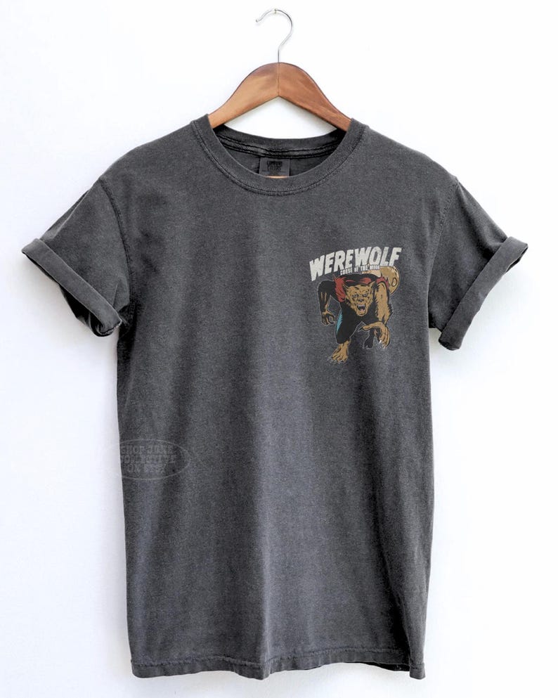 May include: A charcoal gray short-sleeve t-shirt featuring a graphic on the upper left chest. The graphic depicts a brown werewolf with a red cape and the text "WEREWOLF" in white capital letters above the creature. The shirt has a crew neckline.