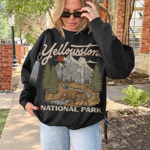 May include: A black long-sleeve sweatshirt featuring a graphic print of Yellowstone National Park. The design includes a brown bison, green pine trees, a winding river, and snow-capped mountains under a red sun, with the text Yellowstone National Park.