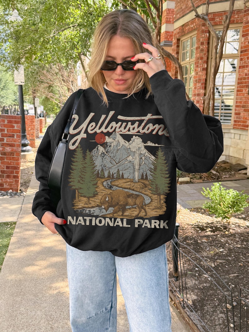May include: A black long-sleeve sweatshirt featuring a graphic print of Yellowstone National Park. The design includes a brown bison, green pine trees, a winding river, and snow-capped mountains under a red sun, with the text Yellowstone National Park.