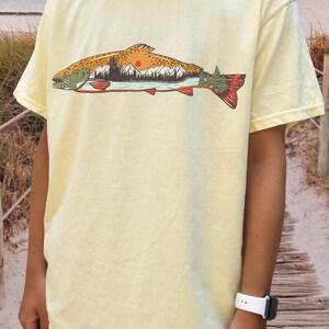 Puede incluir: Camiseta amarillo claro con un dise&ntilde;o de pez. La ilustraci&oacute;n del pez contiene un paisaje con monta&ntilde;as, un lago y un sol poniente. El pez tiene detalles en naranja, rojo y verde.