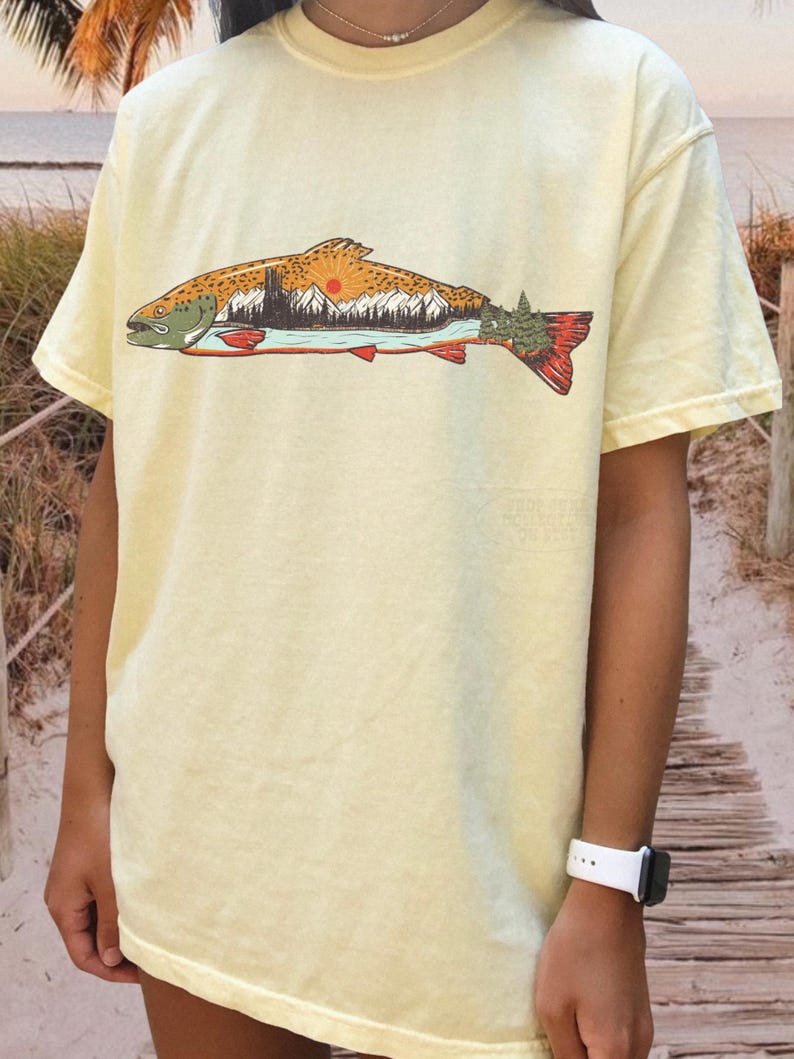 Puede incluir: Camiseta amarillo claro con un dise&ntilde;o de pez. La ilustraci&oacute;n del pez contiene un paisaje con monta&ntilde;as, un lago y un sol poniente. El pez tiene detalles en naranja, rojo y verde.