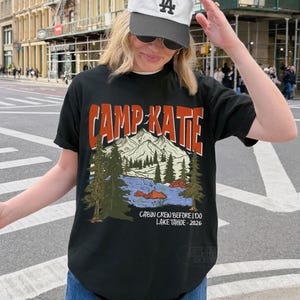 May include: Black t-shirt with the text "CAMP KATIE" in orange and white. The shirt features a mountain and lake graphic with the text "CABIN CREW BEFORE I DO LAKE TAHOE - 2026". The person is wearing a white baseball cap.