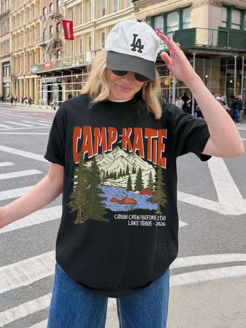 May include: Black t-shirt with the text "CAMP KATIE" in orange and white. The shirt features a mountain and lake graphic with the text "CABIN CREW BEFORE I DO LAKE TAHOE - 2026". The person is wearing a white baseball cap.