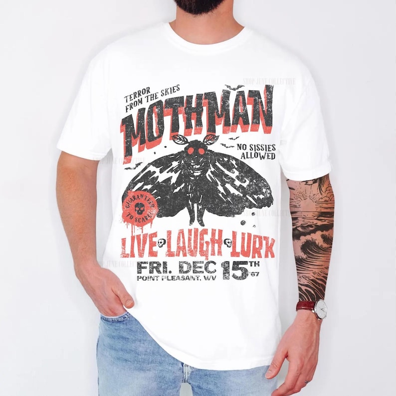 May include: A white t-shirt featuring a black moth graphic with red eyes and the text "TERROR FROM THE SKIES MOTHMAN NO SISSIES ALLOWED GUARANTEED TO SCARE LIVE LAUGH LURK FRI. DEC 15TH 67 POINT PLEASANT, WV" printed in black and red distressed lettering.