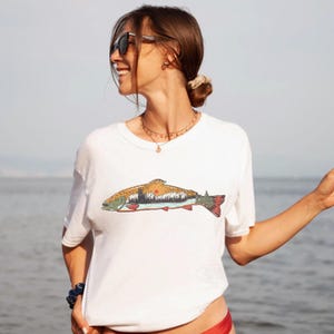 Puede incluir: Camiseta blanca con una colorida ilustraci&oacute;n de un pez con un dise&ntilde;o de monta&ntilde;a y puesta de sol. El pez tiene detalles en naranja, verde y rojo. La camiseta es de estilo casual y de manga corta.