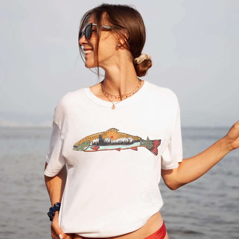 Puede incluir: Camiseta blanca con una colorida ilustraci&oacute;n de un pez con un dise&ntilde;o de monta&ntilde;a y puesta de sol. El pez tiene detalles en naranja, verde y rojo. La camiseta es de estilo casual y de manga corta.