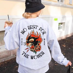 May include: A light gray sweatshirt with a graphic on the back that says "NO REST FOR THE WOMEN" with a panther, flames, and a rose. The sweatshirt is worn with a black bucket hat.
