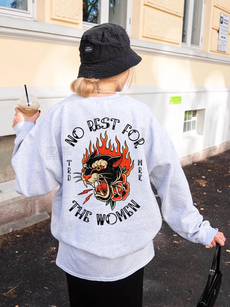 May include: A light gray sweatshirt with a graphic on the back that says "NO REST FOR THE WOMEN" with a panther, flames, and a rose. The sweatshirt is worn with a black bucket hat.