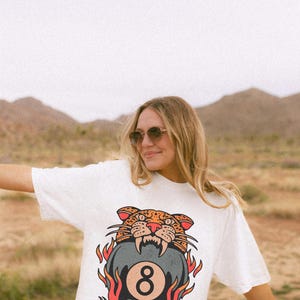 May include: White t-shirt featuring a graphic of a snarling leopard biting a pool 8-ball, surrounded by flames. The design is in shades of orange, red, black, and gray. The shirt is worn by a person in a desert landscape.