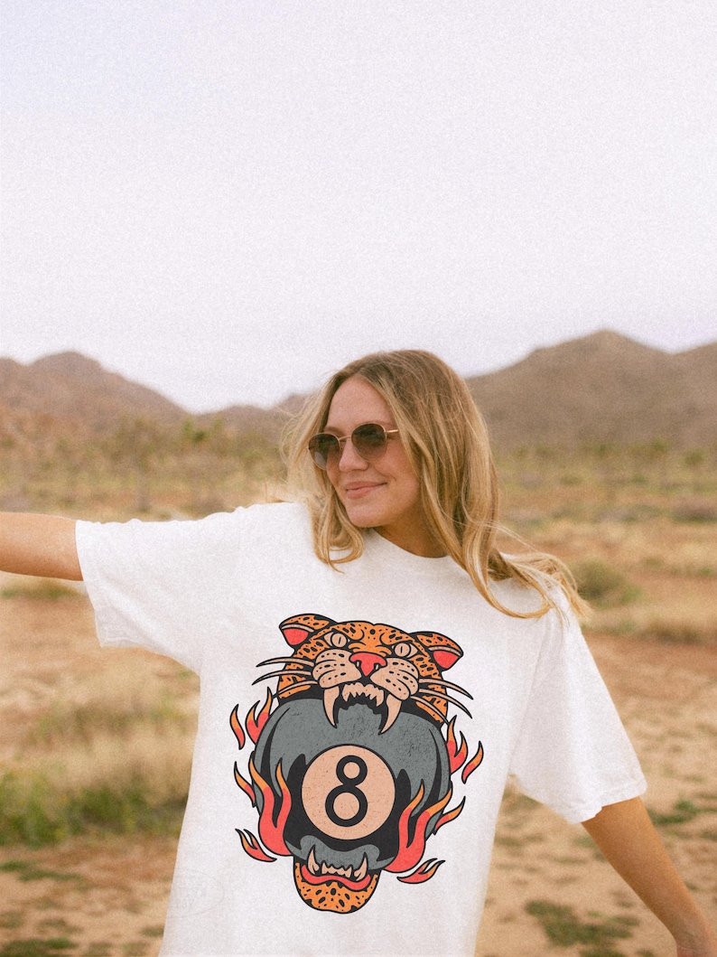 May include: White t-shirt featuring a graphic of a snarling leopard biting a pool 8-ball, surrounded by flames. The design is in shades of orange, red, black, and gray. The shirt is worn by a person in a desert landscape.