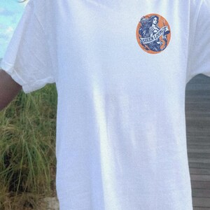 May include: White t-shirt with a graphic of a mermaid with the text "Siren Lounge" on a circular orange background.