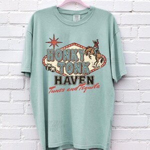 Pode incluir: Uma camiseta verde claro com um design gr&aacute;fico vintage que diz "Honky Tonk Haven". O design inclui um cowboy montando um cavalo e uma estrela vermelha. O texto "Tunes and Tequila" tamb&eacute;m est&aacute; inclu&iacute;do.